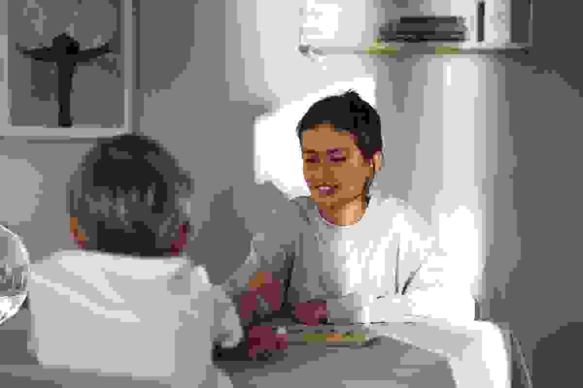 Två barn sitter vid ett bord och spelar ett brädspel i ett solbelyst rum med en tavla och en hylla i bakgrunden